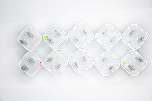Nine white electrical plug adapters are arranged in a diamond pattern on a white background. Each adapter has two flat prongs, and some have small circular markings in green or pink.