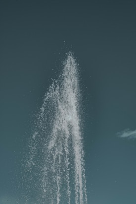 Water shoots upward in a high, powerful stream against a blue-green sky. The spray creates a dynamic pattern as it arcs and falls back down.
