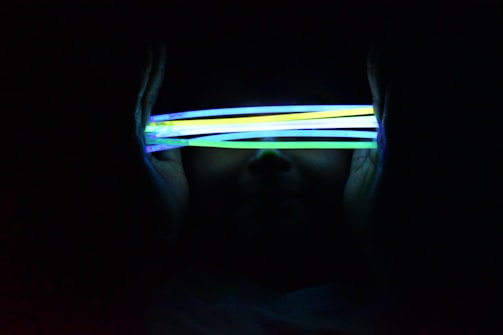 Several brightly colored glow sticks are held horizontally in front of a face, partially obscuring it. The glow sticks emit vivid colors such as yellow, green, and blue, creating a contrast against the dark background.