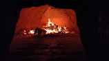 A warm, glowing fireplace with burning logs inside a brick or stone structure. The flames flicker, casting an orange and red hue throughout the enclosed space, creating a cozy ambiance.