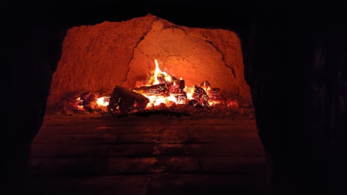 A warm, glowing fireplace with burning logs inside a brick or stone structure. The flames flicker, casting an orange and red hue throughout the enclosed space, creating a cozy ambiance.
