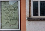A piece of paper with handwritten text in German is taped to a window. The surrounding area includes parts of a building exterior with orange and beige walls and wooden window frames.