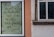 A piece of paper with handwritten text in German is taped to a window. The surrounding area includes parts of a building exterior with orange and beige walls and wooden window frames.