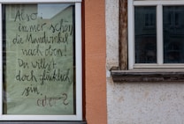 A piece of paper with handwritten text in German is taped to a window. The surrounding area includes parts of a building exterior with orange and beige walls and wooden window frames.