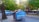 A blue dumpster with weathered paint and the words 'Fatin Soe Lixo' is situated on a street adjacent to a parked vehicle labeled 'HW Lifestyle.' The setting includes lush green trees providing shade over the street, while a pedestrian area with benches is visible in the background.