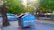 A blue dumpster with weathered paint and the words 'Fatin Soe Lixo' is situated on a street adjacent to a parked vehicle labeled 'HW Lifestyle.' The setting includes lush green trees providing shade over the street, while a pedestrian area with benches is visible in the background.