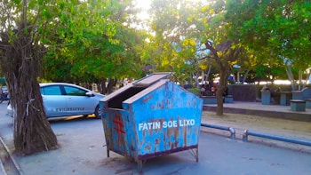 A blue dumpster with weathered paint and the words 'Fatin Soe Lixo' is situated on a street adjacent to a parked vehicle labeled 'HW Lifestyle.' The setting includes lush green trees providing shade over the street, while a pedestrian area with benches is visible in the background.