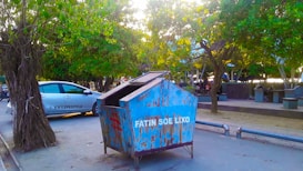 A blue dumpster with weathered paint and the words 'Fatin Soe Lixo' is situated on a street adjacent to a parked vehicle labeled 'HW Lifestyle.' The setting includes lush green trees providing shade over the street, while a pedestrian area with benches is visible in the background.