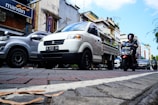 A busy urban street scene with various vehicles, including a white pickup truck and a motorcycle, moving along a paved road. The background features buildings with signage, including a Mandiri bank sign, and there are other cars visible. Pedestrians are likely present, although not the central focus.