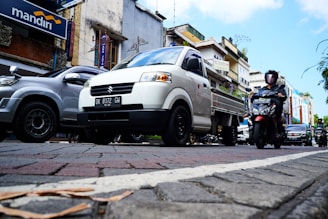 A busy urban street scene with various vehicles, including a white pickup truck and a motorcycle, moving along a paved road. The background features buildings with signage, including a Mandiri bank sign, and there are other cars visible. Pedestrians are likely present, although not the central focus.