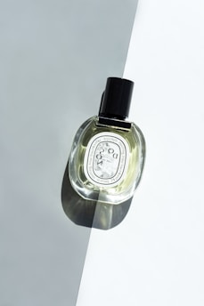 A glass bottle of perfume with a black cap is placed on a surface divided into light and dark shades. The bottle casts a shadow, and the label features text and design elements.