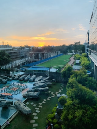 A wide view of an outdoor area featuring a landscaped garden with large rocks arranged artistically. A pathway made of square stones meanders through the grass, surrounded by neatly trimmed bushes. In the background, a sports field extends into the distance, bordered by a blue fence. The setting sun on the horizon casts a warm glow across the scene, with partially cloudy skies overhead.