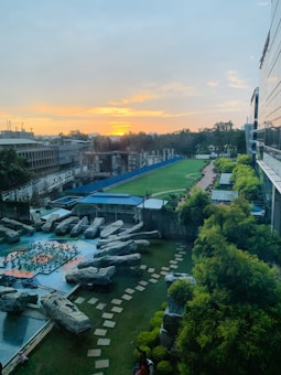 A wide view of an outdoor area featuring a landscaped garden with large rocks arranged artistically. A pathway made of square stones meanders through the grass, surrounded by neatly trimmed bushes. In the background, a sports field extends into the distance, bordered by a blue fence. The setting sun on the horizon casts a warm glow across the scene, with partially cloudy skies overhead.