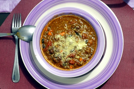 A bowl of hearty stew is served on a purple and white striped plate with a spoon and fork placed on a burgundy placemat. The stew contains visible beans, diced vegetables, and is topped with grated cheese.