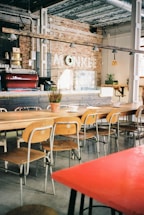 A cozy cafe interior with a long wooden communal table surrounded by mismatched chairs. The space is decorated with exposed brick walls and industrial-style metal pipes. A coffee bar features an espresso machine and bar stools, while hanging pendant lights provide soft lighting.