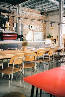 A cozy cafe interior with a long wooden communal table surrounded by mismatched chairs. The space is decorated with exposed brick walls and industrial-style metal pipes. A coffee bar features an espresso machine and bar stools, while hanging pendant lights provide soft lighting.