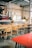 A cozy cafe interior with a long wooden communal table surrounded by mismatched chairs. The space is decorated with exposed brick walls and industrial-style metal pipes. A coffee bar features an espresso machine and bar stools, while hanging pendant lights provide soft lighting.