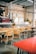 A cozy cafe interior with a long wooden communal table surrounded by mismatched chairs. The space is decorated with exposed brick walls and industrial-style metal pipes. A coffee bar features an espresso machine and bar stools, while hanging pendant lights provide soft lighting.
