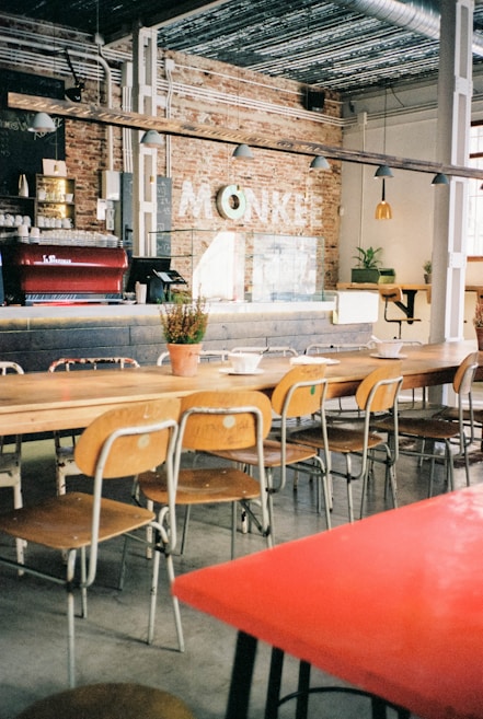 A cozy cafe interior with a long wooden communal table surrounded by mismatched chairs. The space is decorated with exposed brick walls and industrial-style metal pipes. A coffee bar features an espresso machine and bar stools, while hanging pendant lights provide soft lighting.