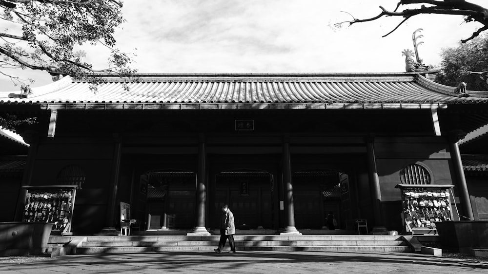 grayscale photo of man walking on sidewalk near building