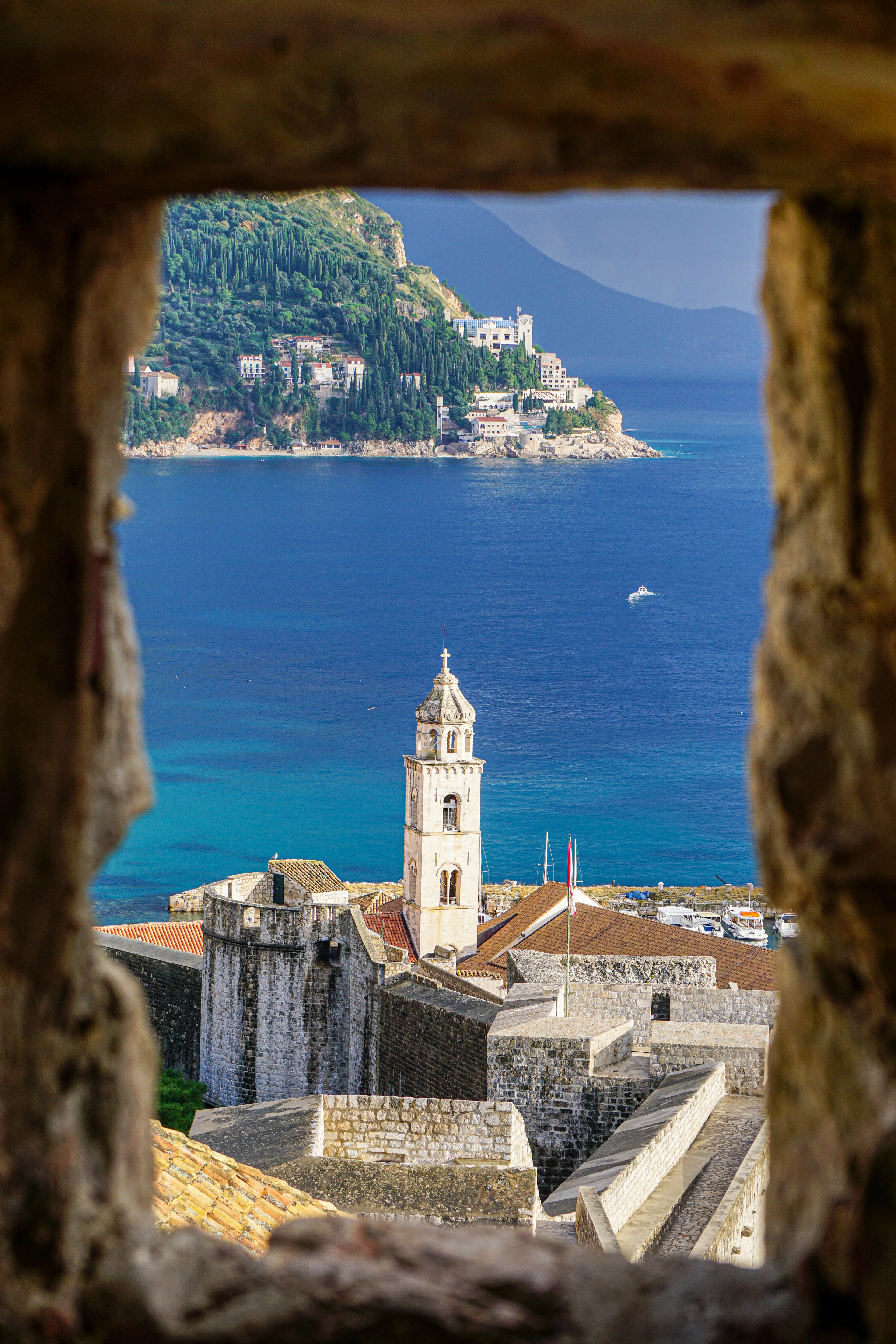 A stone loophole in Dubrovnik's city walls frames the bell tower, red rooftops and Lokrum island across the sea.
