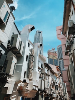 Multiple buildings with external air conditioning units and large ductwork dominate the scene. In the background, tall skyscrapers under construction are visible against a clear blue sky. The view is from a narrow alley with buildings on both sides.