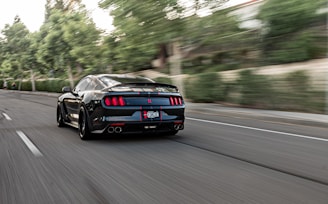 A sleek black sports car is driving at high speed down a road lined with trees. The background is blurred, indicating motion and speed. The car has distinctive rear lights and a visible license plate.