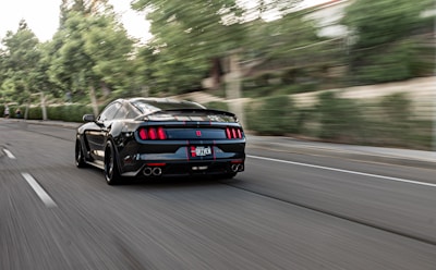 A sleek black sports car is driving at high speed down a road lined with trees. The background is blurred, indicating motion and speed. The car has distinctive rear lights and a visible license plate.