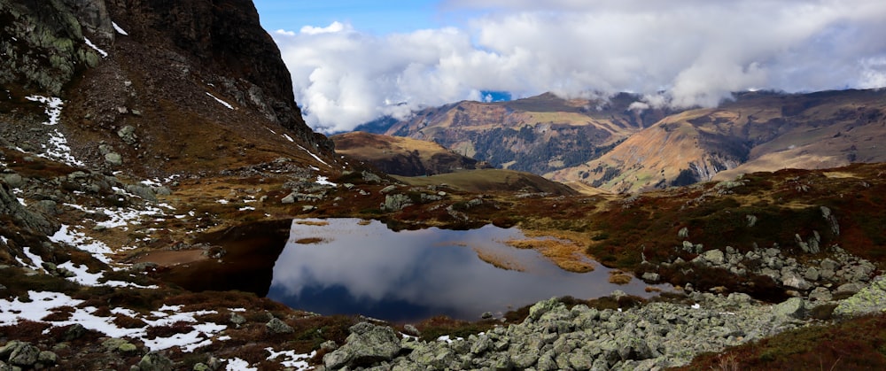 brown rocky mountain beside lake under white clouds and blue sky during daytime