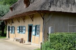 A rustic, thatched-roof cottage featuring three blue and white shuttered windows and a door. The structure is accented with wooden beams, and a bench rests against the wall. Dense greenery and trees surround the building, enhancing its quaint, rural charm.