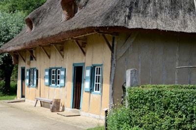 A rustic, thatched-roof cottage featuring three blue and white shuttered windows and a door. The structure is accented with wooden beams, and a bench rests against the wall. Dense greenery and trees surround the building, enhancing its quaint, rural charm.
