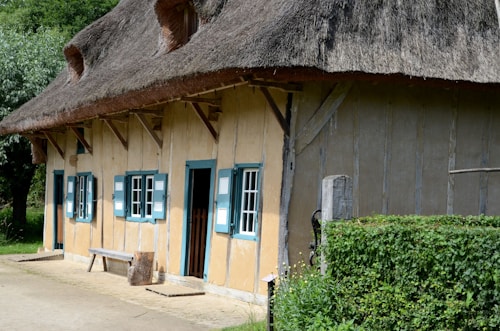 A rustic, thatched-roof cottage featuring three blue and white shuttered windows and a door. The structure is accented with wooden beams, and a bench rests against the wall. Dense greenery and trees surround the building, enhancing its quaint, rural charm.