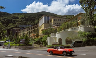 A bright red convertible car drives along a road in front of a large, elegant building set against a backdrop of lush green mountains. The building features light-colored walls and numerous windows, accompanied by landscaped gardens and trees. In the distance, a clear blue sky with fluffy white clouds can be seen, enhancing the picturesque mountain setting. Vehicles, including a van, are parked nearby, while signage in a foreign language is visible at the roadside.