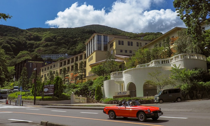 A bright red convertible car drives along a road in front of a large, elegant building set against a backdrop of lush green mountains. The building features light-colored walls and numerous windows, accompanied by landscaped gardens and trees. In the distance, a clear blue sky with fluffy white clouds can be seen, enhancing the picturesque mountain setting. Vehicles, including a van, are parked nearby, while signage in a foreign language is visible at the roadside.
