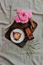 A cup of coffee with latte art on a saucer surrounded by nuts and berries. Pink flowers are situated at the top left on the tray along with a dark chocolate bar and some cinnamon sticks. The composition rests on a brown wicker pattern tray against a beige fabric background.
