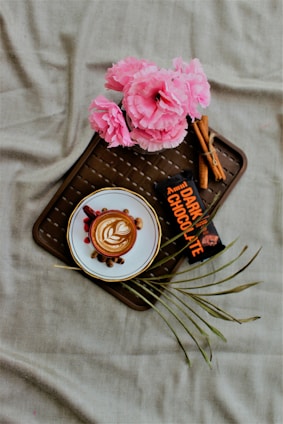 A cup of coffee with latte art on a saucer surrounded by nuts and berries. Pink flowers are situated at the top left on the tray along with a dark chocolate bar and some cinnamon sticks. The composition rests on a brown wicker pattern tray against a beige fabric background.