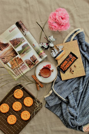 An arrangement of various items on a beige fabric surface includes an open magazine displaying travel photography, a pink artificial flower, and a piece of dark chocolate with partially open foil. There is a small decorative ship model, honeycomb filled cup on a saucer with cinnamon sticks and cardamom pods nearby, a blue fabric draped next to a notebook, and a tray with round crackers.