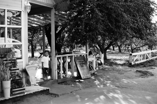 A black-and-white image of a rural outdoor setting with a small wooden porch area. A child in white clothing stands near the entrance, looking towards the camera. To the side, a couple sits at a table engaged in conversation, surrounded by trees. A wooden fence runs along the background, and various potted plants and a sign are visible.