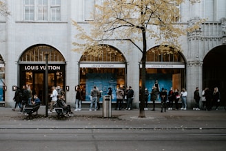 A group of people is gathered outside a Louis Vuitton store. The storefront features large arches and elegant window displays with blue backgrounds. A tree with yellow autumn leaves stands in front of the store, and a few people are sitting on a bench nearby. The atmosphere is urban with tram tracks in the foreground.