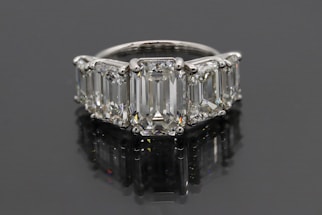 A luxurious ring featuring five large, rectangular emerald-cut diamonds set in a row. The diamonds are mounted in a shiny metal band, likely platinum, and reflect a brilliant shine against a dark background.