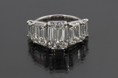 A luxurious ring featuring five large, rectangular emerald-cut diamonds set in a row. The diamonds are mounted in a shiny metal band, likely platinum, and reflect a brilliant shine against a dark background.