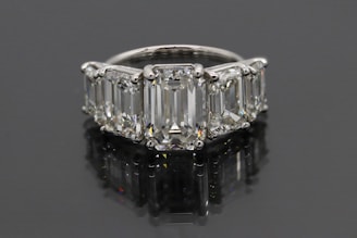 A luxurious ring featuring five large, rectangular emerald-cut diamonds set in a row. The diamonds are mounted in a shiny metal band, likely platinum, and reflect a brilliant shine against a dark background.