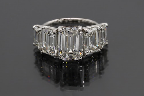 A luxurious ring featuring five large, rectangular emerald-cut diamonds set in a row. The diamonds are mounted in a shiny metal band, likely platinum, and reflect a brilliant shine against a dark background.