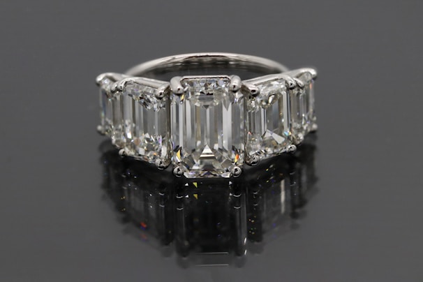 A luxurious ring featuring five large, rectangular emerald-cut diamonds set in a row. The diamonds are mounted in a shiny metal band, likely platinum, and reflect a brilliant shine against a dark background.