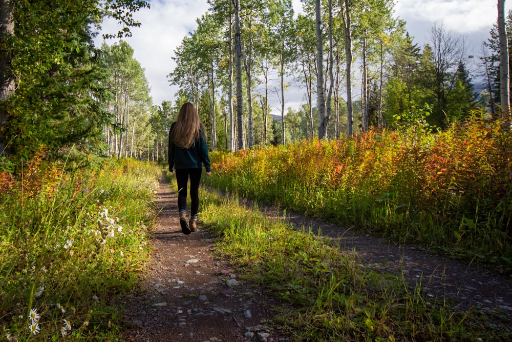 Donna in giacca nera che cammina sul sentiero tra l'erba verde e gli alberi durante il giorno