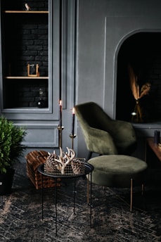 A cozy and stylish interior setting featuring a small black round table with decorative antlers and pinecones in a wire basket, flanked by two lit tall black candles. In the background, a plush olive-green chair sits beside a dark fireplace. The wall has built-in shelves with small decorative items and a textured black-brick background. A lush green plant is positioned on the floor nearby, enhancing the earthy feel of the room.