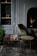 A cozy and stylish interior setting featuring a small black round table with decorative antlers and pinecones in a wire basket, flanked by two lit tall black candles. In the background, a plush olive-green chair sits beside a dark fireplace. The wall has built-in shelves with small decorative items and a textured black-brick background. A lush green plant is positioned on the floor nearby, enhancing the earthy feel of the room.
