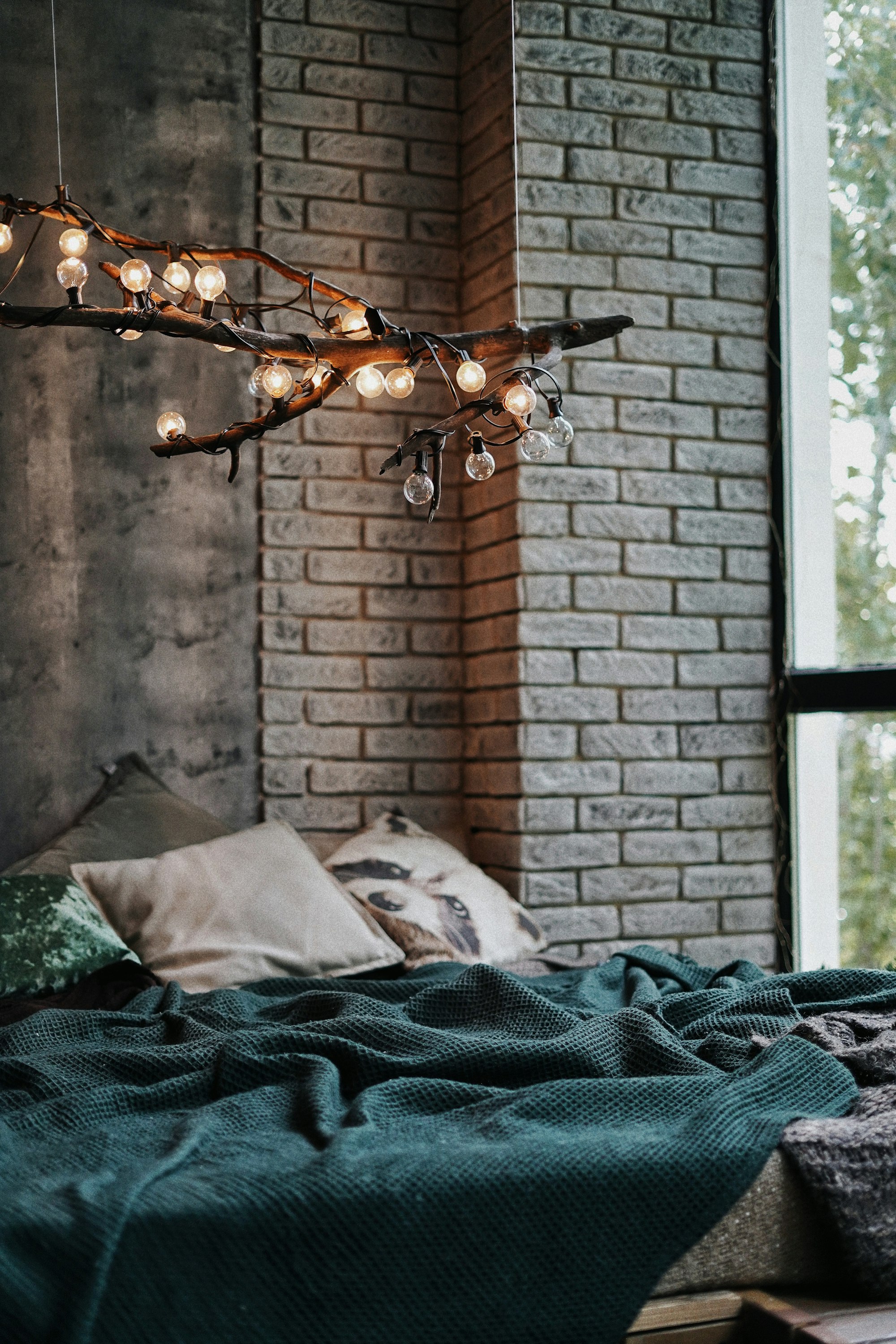 Fairy lights draped across a large stick to create a moody atmosphere. The bedroom has dark white brick walls, large windows, and a bed. The bed has a deep blue color throw to add a pop of color.