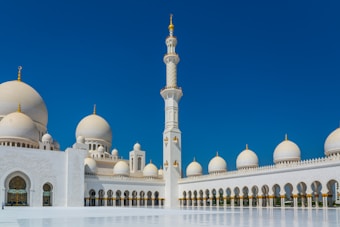 A grand mosque with large white domes and a prominent minaret against a clear blue sky. The architectural design is intricate with detailed carvings and gold accents.