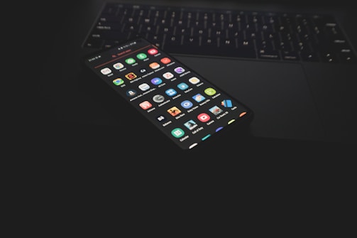 A smartphone displaying multiple apps on its screen is placed on a partially visible laptop keyboard. The scene is dimly lit, emphasizing the colorful icons on the phone screen against the dark surroundings.